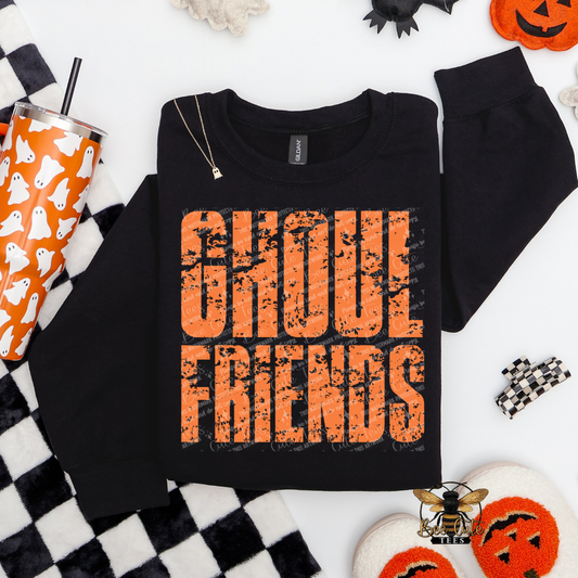 Black crewneck sweatshirt featuring a distressed orange and black “Ghoul Friends” Halloween graphic, styled with an orange ghost tumbler, checkered blanket, pumpkin slippers, and Halloween decorations.