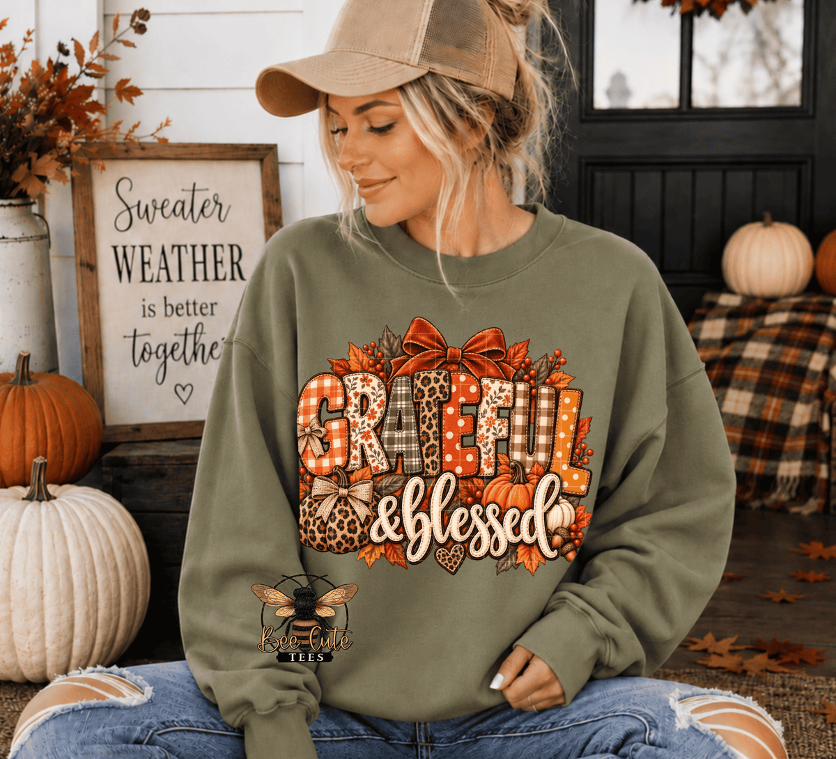 A woman wearing an olive green crewneck sweatshirt with a large 'Grateful & Blessed' autumn graphic sits in a decorated fall porch setting with pumpkins and a sign.