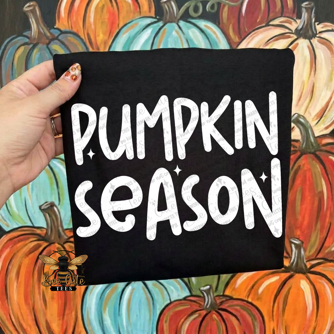 A hand holds a black folded t-shirt featuring the words “PUMPKIN SEASON” in large white decorative letters, set against a colorful painted background of pumpkins.