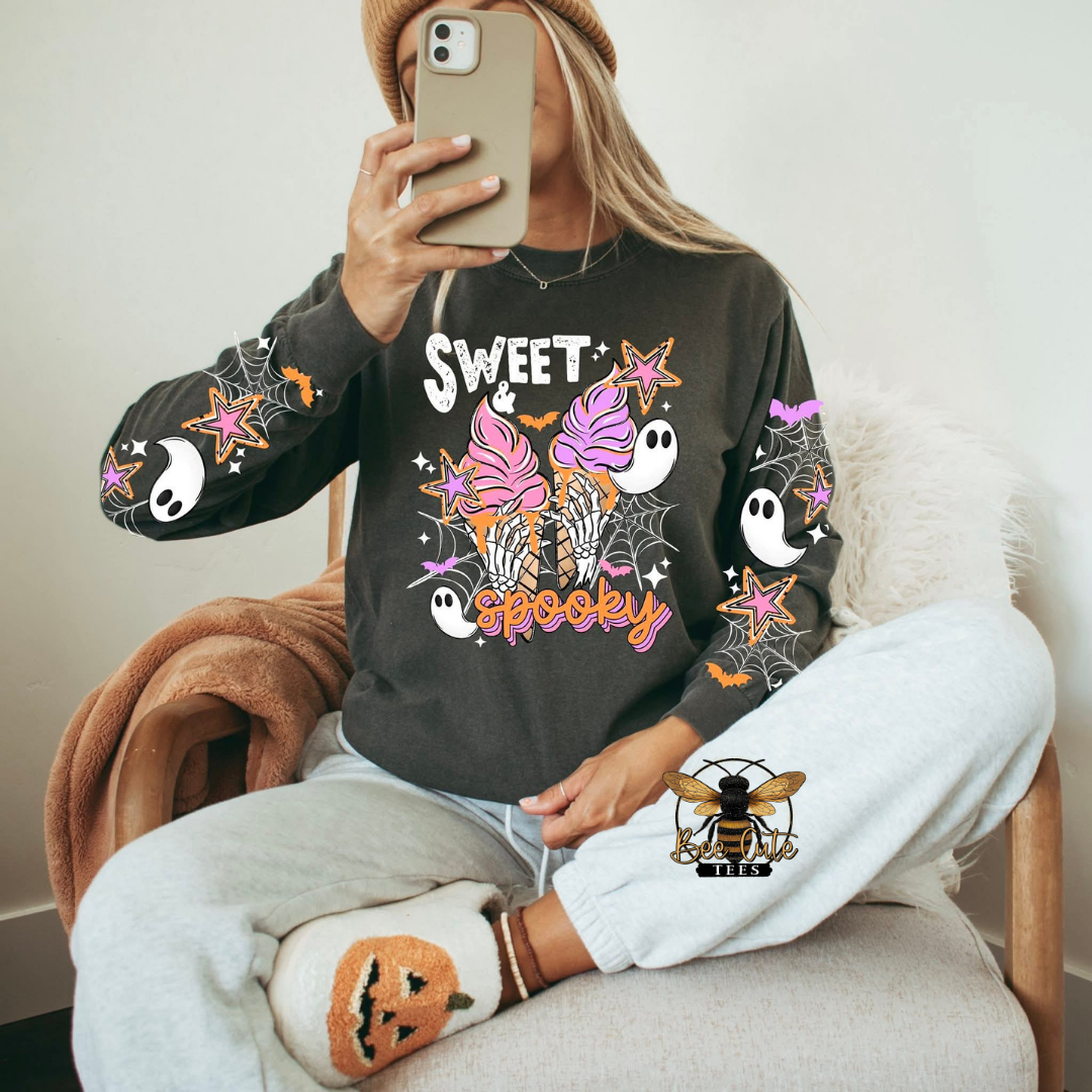 A woman takes a mirror selfie wearing a dark gray long-sleeve sweatshirt with a colorful 'Sweet Spooky' Halloween graphic, paired with light gray sweatpants and a brown beanie.