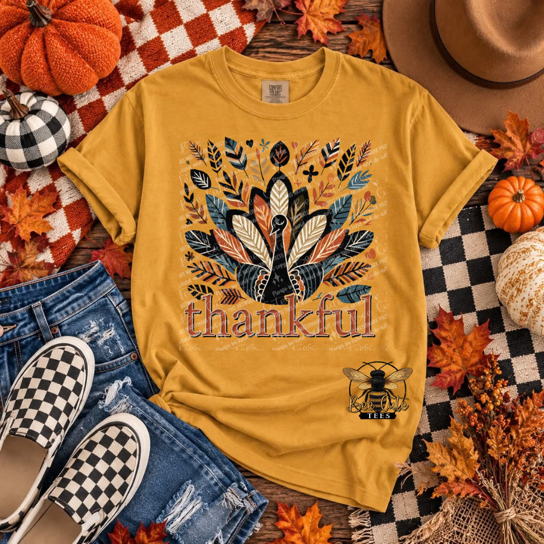 A mustard yellow graphic t-shirt featuring a stylized turkey surrounded by autumn leaves and the word 'thankful' in distressed lettering. The shirt is presented in a flat-lay arrangement with jeans, checkered shoes, pumpkins, a hat, and fall leaves on a rustic wooden background.