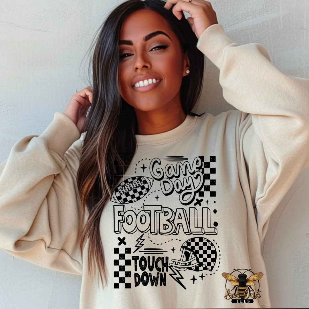 A woman wearing a beige crewneck sweatshirt with a black and white football graphic that reads 'Game Day FOOTBALL' and 'TOUCH DOWN', along with a bee logo, against a light gray wall.