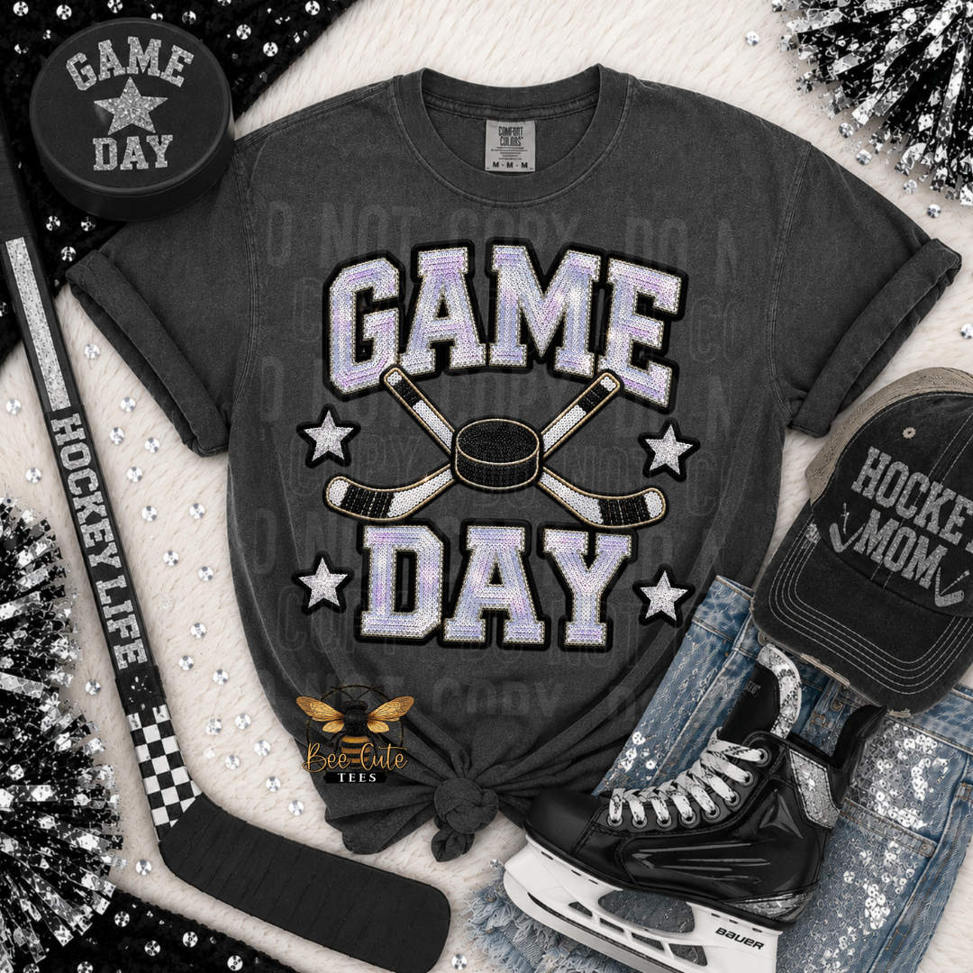A dark gray graphic t-shirt with a 'GAME DAY' hockey design, styled in a flat-lay arrangement with a hockey puck, stick, skates, jeans, and a cap.