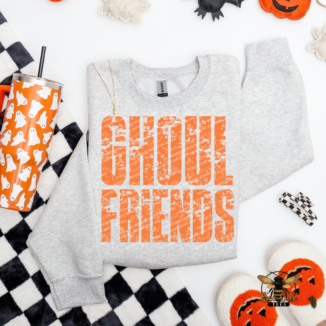Heather gray crewneck sweatshirt featuring a distressed orange “Ghoul Friends” Halloween graphic, styled with an orange ghost tumbler, black-and-white checkered blanket, pumpkin slippers, and festive Halloween accents.