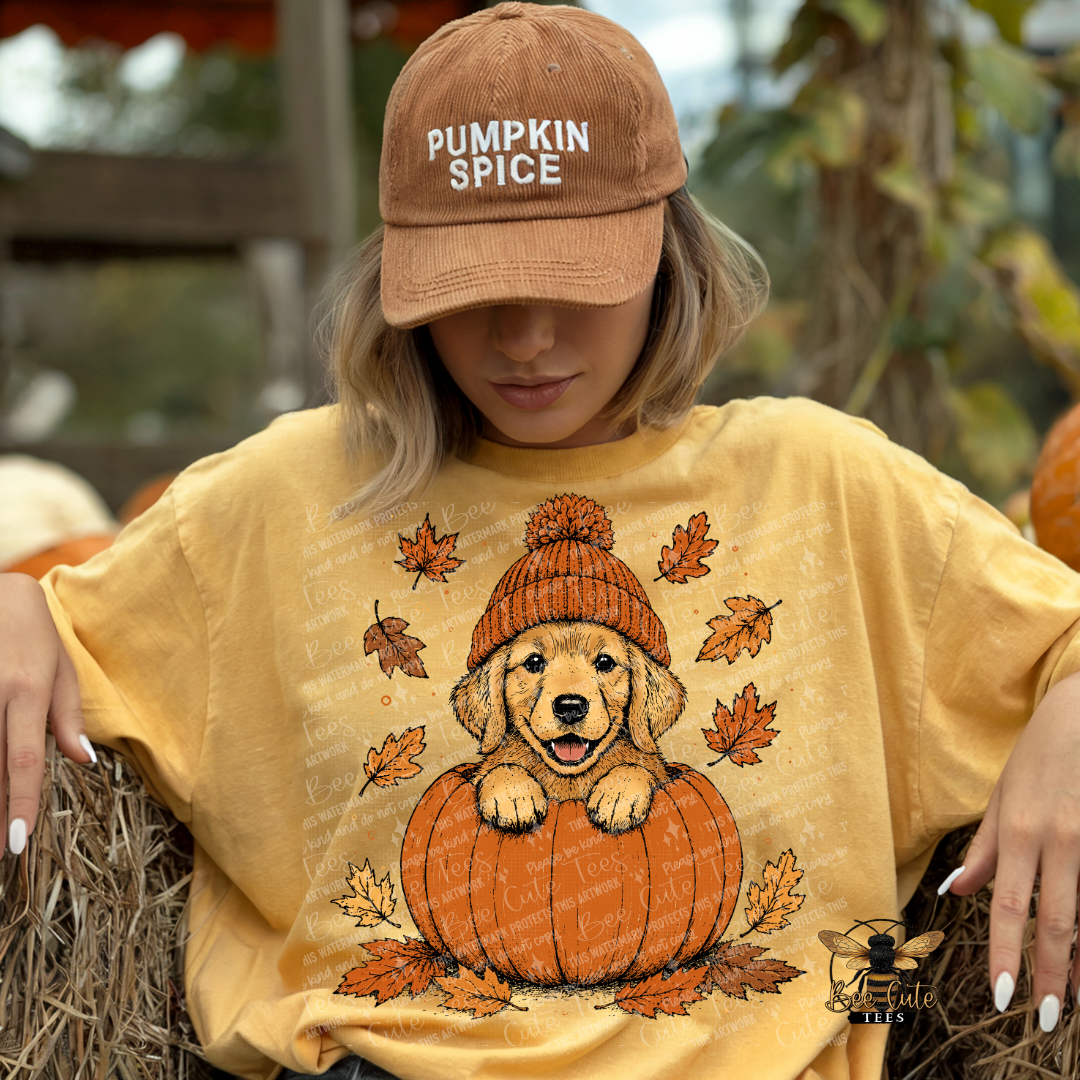 A woman wearing a tan baseball cap that says 'PUMPKIN SPICE' and a yellow graphic t-shirt featuring a golden retriever puppy sitting on a pumpkin with autumn leaves. She is posed on a hay bale in an outdoor fall-themed setting.
