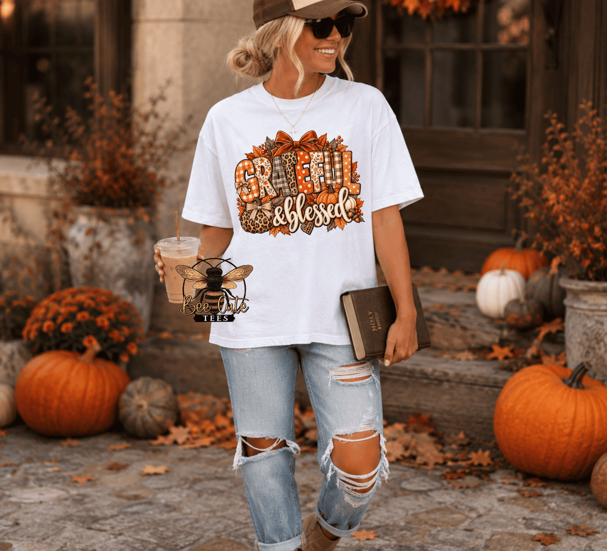 A woman wearing a white graphic t-shirt with a 'Grateful & Blessed' autumn design, paired with distressed light blue jeans, standing in an outdoor fall-themed setting with pumpkins and autumn leaves.