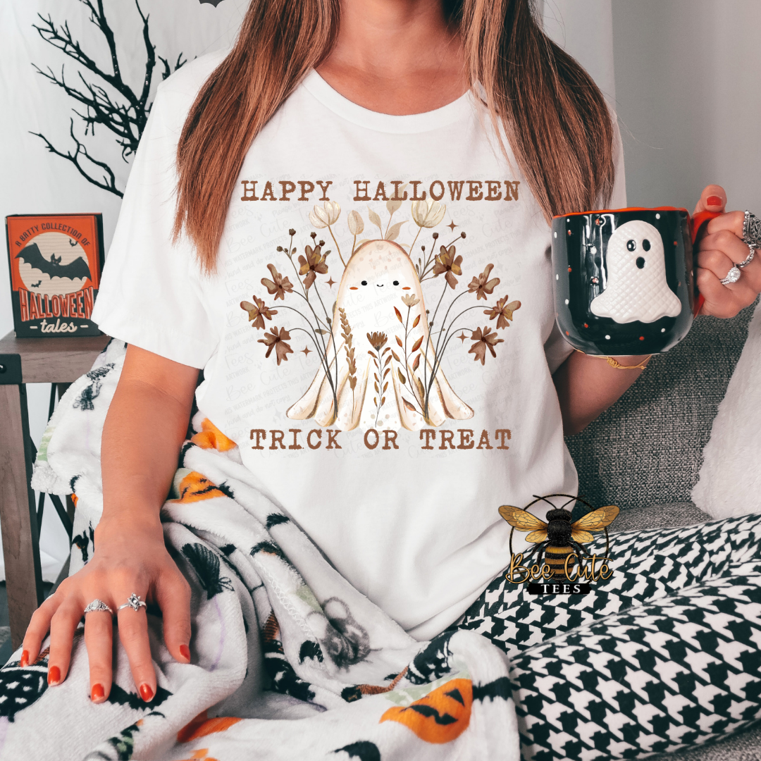A woman wearing a white graphic t-shirt with a cute ghost illustration and the text 'HAPPY HALLOWEEN TRICK OR TREAT'. She is holding a black mug with a white ghost, sitting on a couch with a patterned blanket.