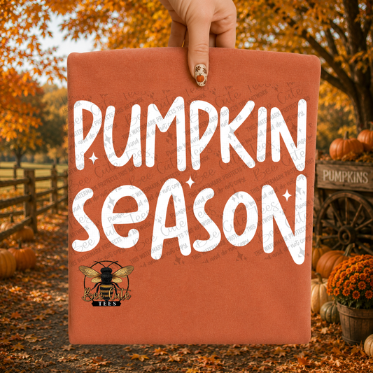A hand holds a rust-orange folded t-shirt-style display with large white text reading “PUMPKIN SEASON” and a small bee logo near the bottom. The background is an autumn outdoor scene with pumpkins, fall leaves, trees, and a fence.