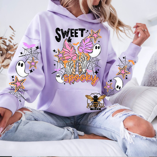 A woman wearing a lavender purple hoodie with a colorful 'Sweet & Spooky' Halloween graphic, paired with ripped light-wash jeans, sitting on a white couch.