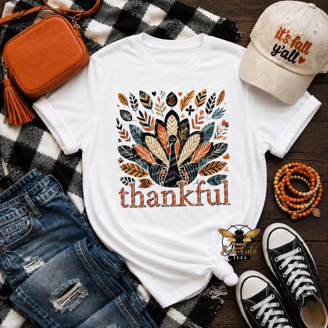 A white graphic T-shirt laid flat on a black-and-white plaid blanket with autumn-themed styling props, including a tan crossbody bag, distressed blue jeans, a beige 'it's fall y'all' cap, wooden bead bracelets, and black canvas sneakers. The shirt features a turkey made from layered leaves in orange, tan, brown, and dark gray, with the word 'thankful' below in orange.