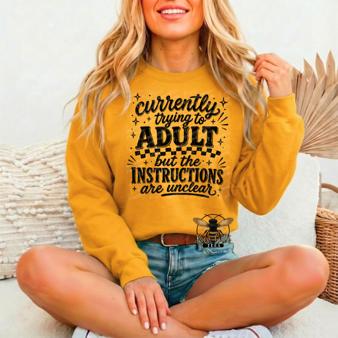 A woman sitting cross-legged wearing a mustard yellow sweatshirt with a black graphic text that reads 'currently trying to ADULT but the INSTRUCTIONS are unclear'.