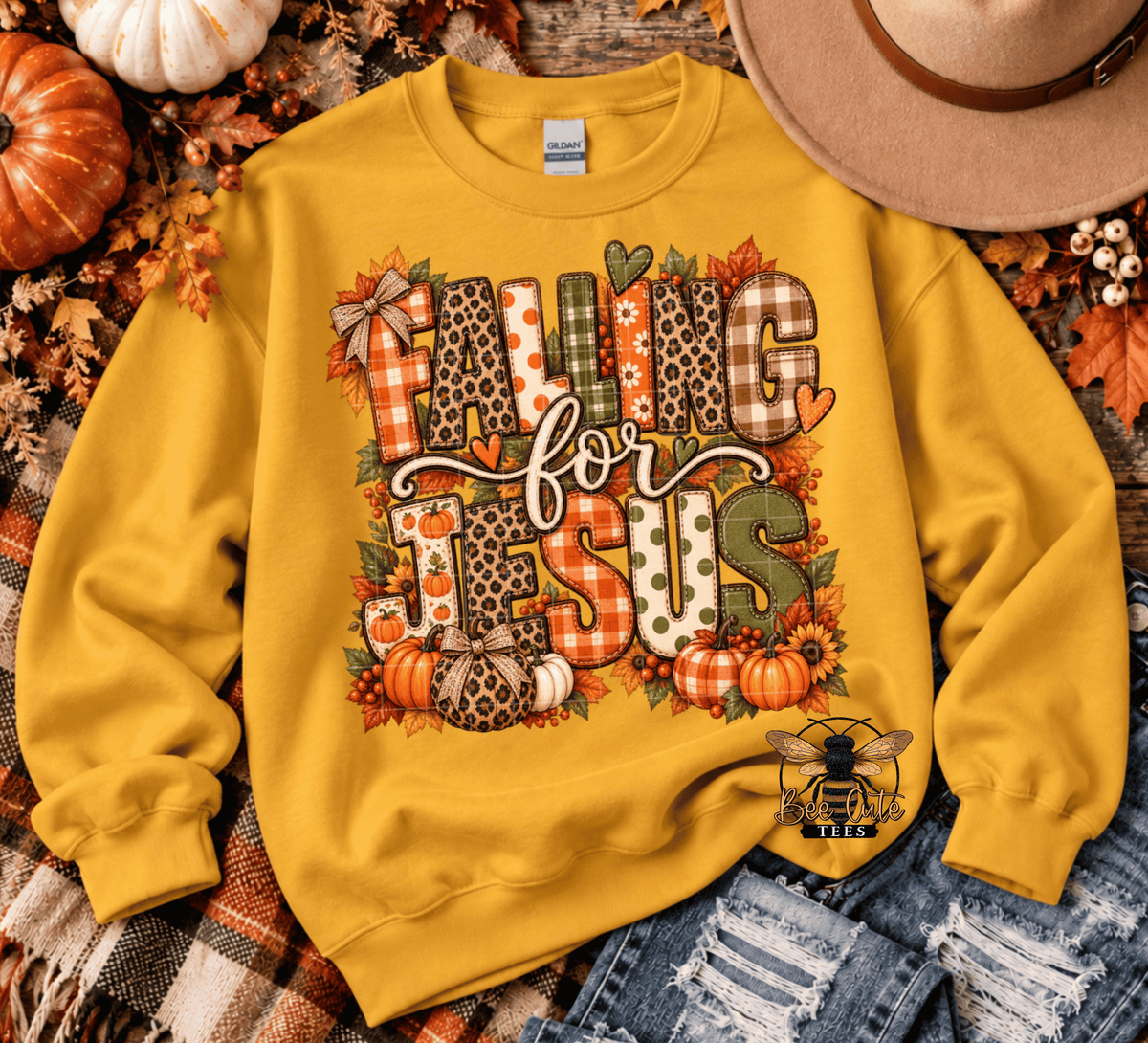 A yellow crewneck sweatshirt with a 'FALLING FOR JESUS' autumn-themed graphic, styled in a flat lay with fall leaves, pumpkins, a hat, and denim jeans.