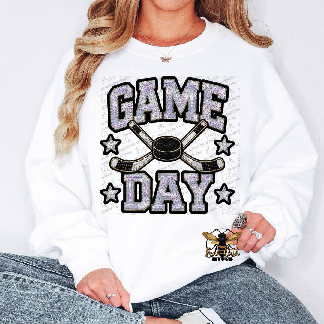 A woman wearing a white crewneck sweatshirt with a 'GAME DAY' hockey graphic featuring crossed hockey sticks, a puck, and stars. The graphic has a glittery silver fill and black outlines.