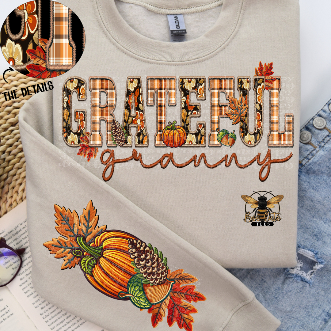 A folded beige crewneck sweatshirt displayed on a flat-lay surface with denim, glasses, and a woven mat. The sweatshirt features a large 'GRATEFUL granny' graphic with autumn-themed fillings, a pumpkin and acorn illustration on the sleeve, and a circular inset showing detailed embroidery.