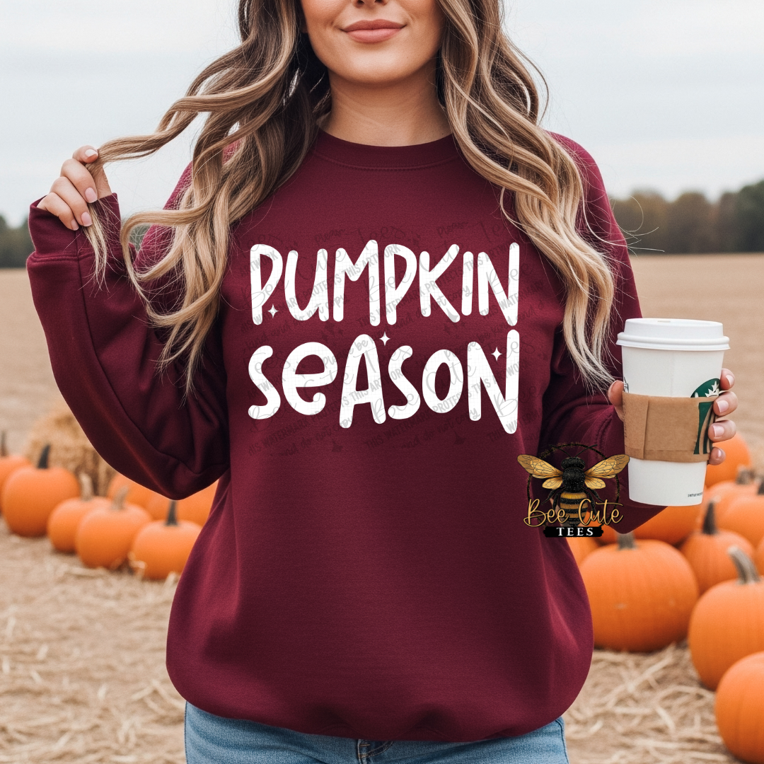 A woman in a maroon sweatshirt with 'PUMPKIN SEASON' printed in white on the front, standing in a pumpkin field and holding a coffee cup. A Bee Cute Tees logo is visible on the lower right of the sweatshirt.