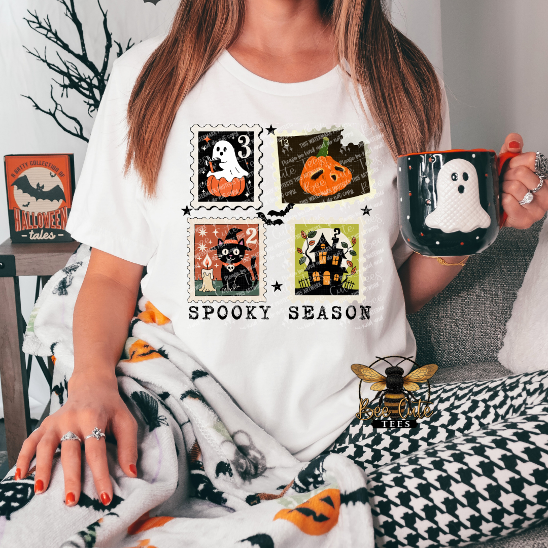 A woman wearing a white graphic t-shirt with a 'Spooky Season' Halloween print, holding a black mug while sitting on a couch under a blanket.
