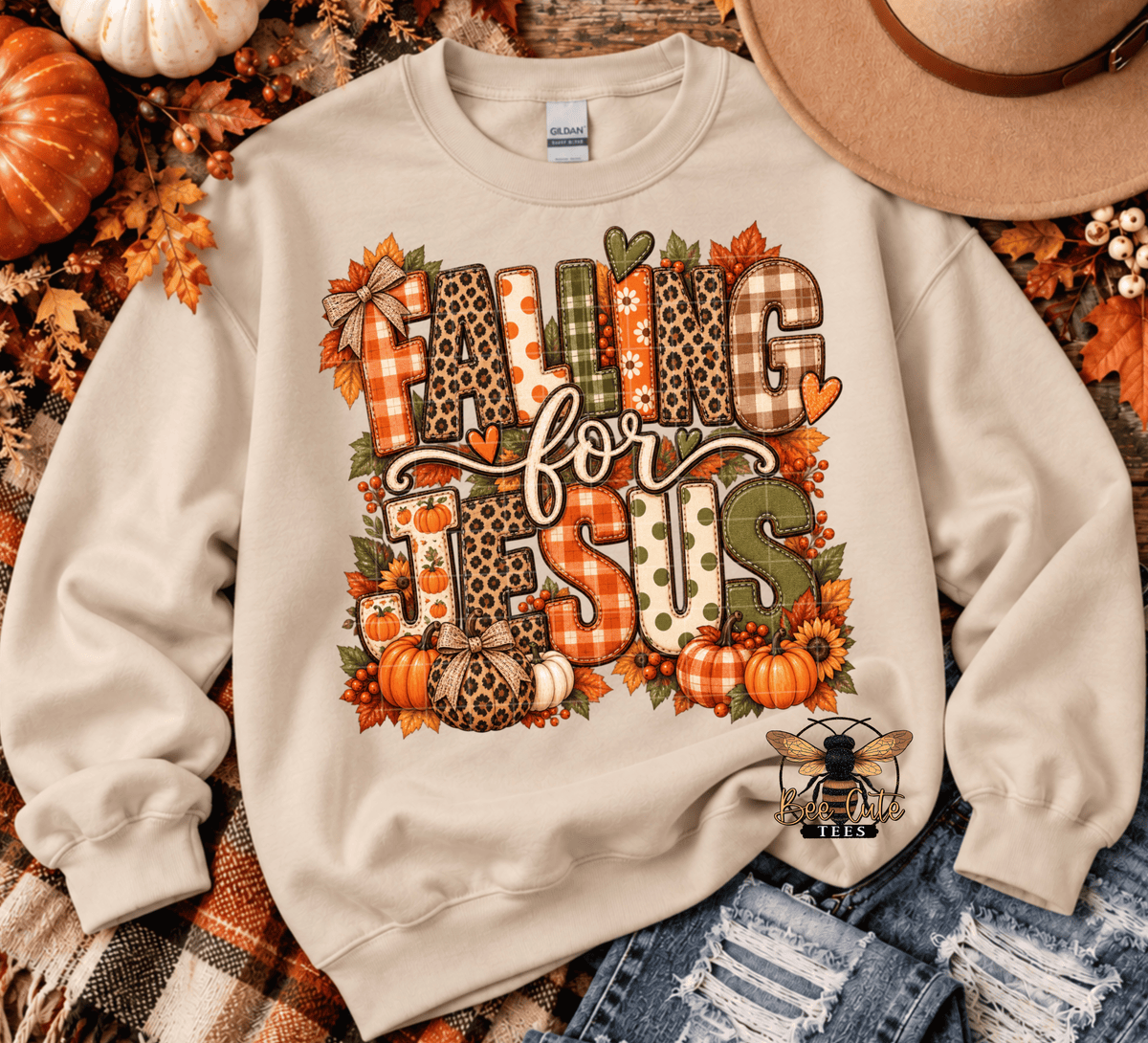 A cream-colored crewneck sweatshirt with a large 'FALLING FOR JESUS' graphic featuring autumn-themed patterns like leopard print, gingham, and plaid, surrounded by pumpkins, leaves, and sunflowers. The sweatshirt is styled in a flat lay with distressed denim, a straw hat, and fall decorations.