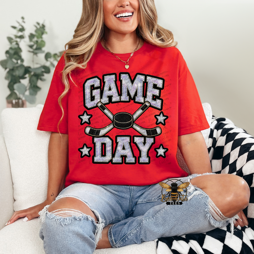 A woman sitting on a white couch wearing a bright red graphic t-shirt that reads 'GAME DAY' in distressed white letters with black outlines, featuring crossed hockey sticks and a hockey puck in the center. She is wearing ripped light-wash blue jeans and a black-and-white patterned throw blanket.