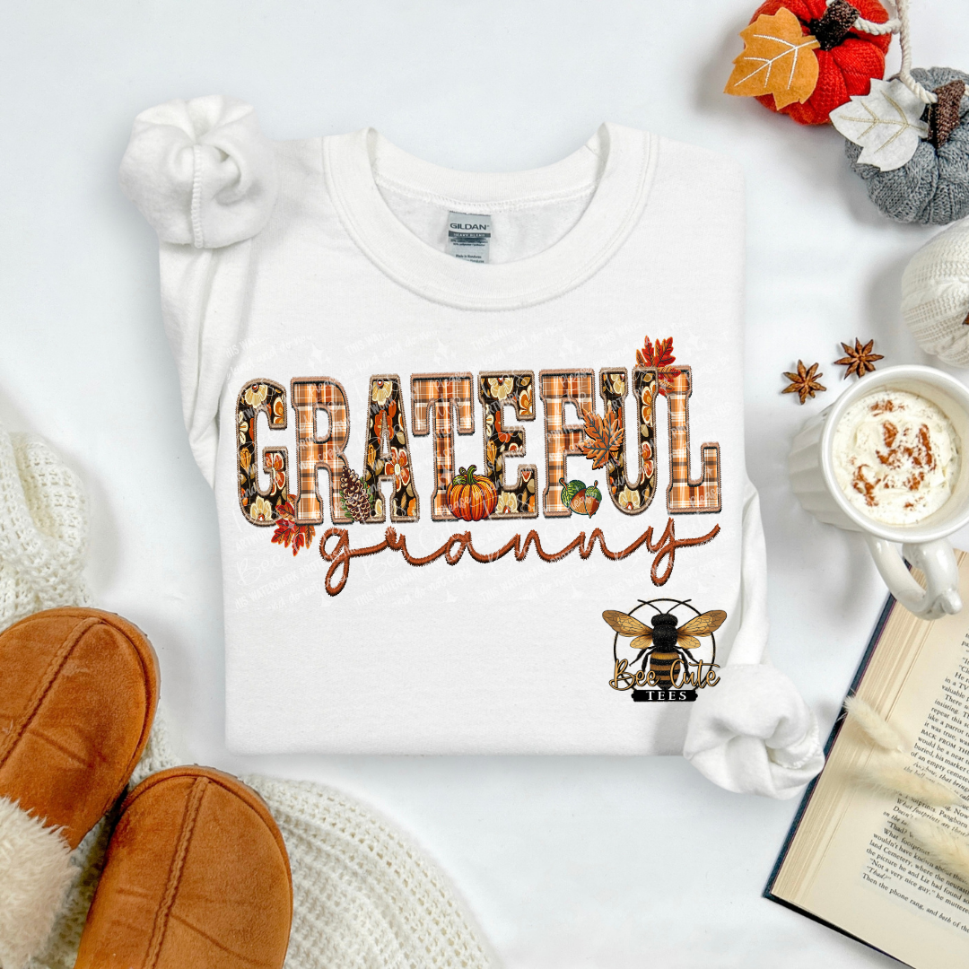 A white long-sleeve t-shirt laid flat on a white surface, featuring a decorative 'GRATEFUL granny' graphic with autumn-themed lettering and a bee logo. The shirt is styled with cozy props including tan slippers, a mug of hot cocoa, an open book, and fall leaves.