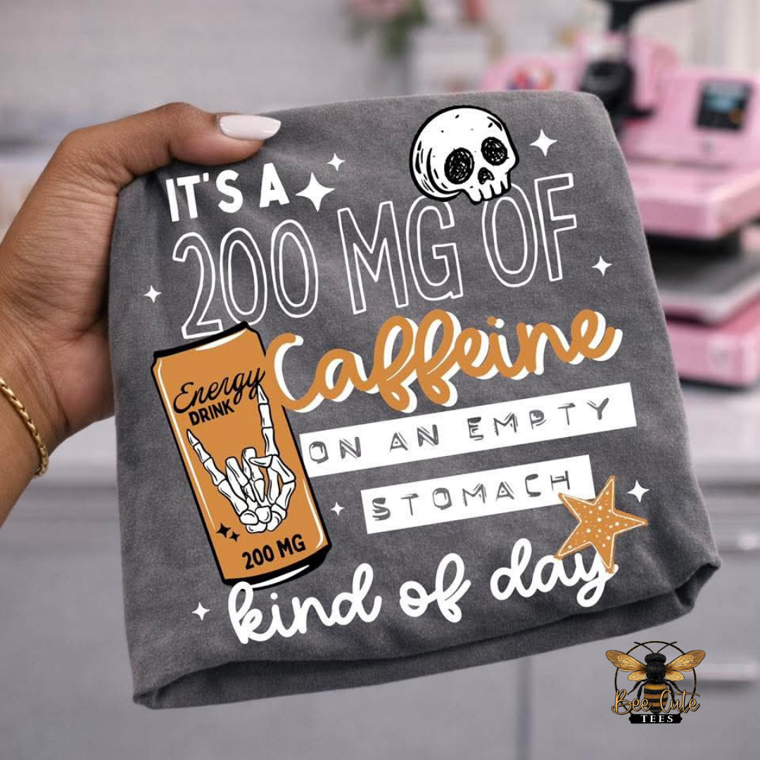 A hand holds a dark gray t-shirt featuring a humorous graphic that reads 'IT'S A 200 MG OF Caffeine ON AN EMPTY STOMACH kind of day' with a cartoon skull and an orange energy drink can illustration.