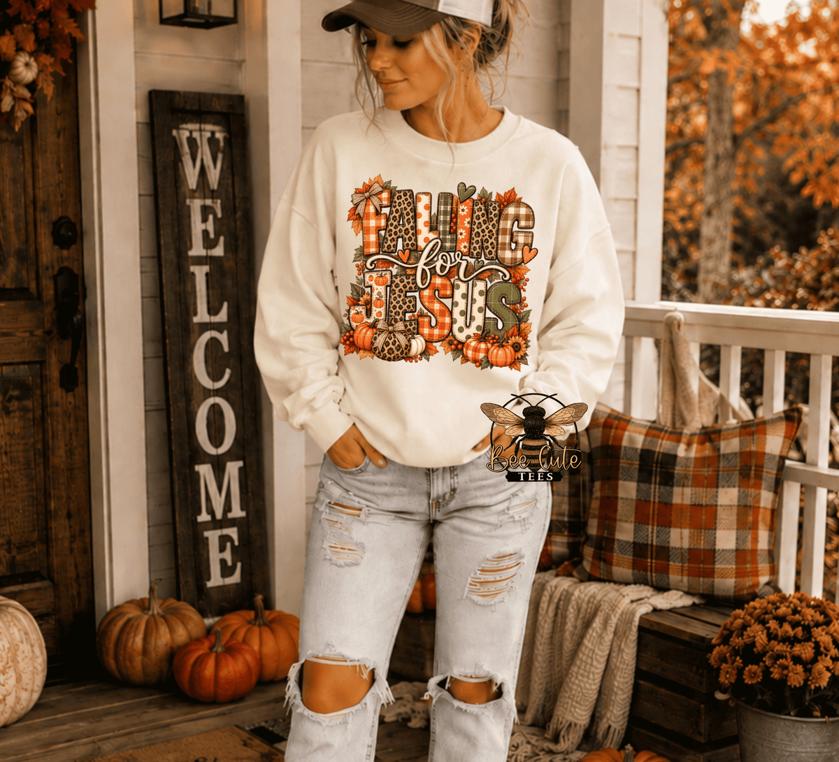 A woman stands on a fall-themed porch wearing a cream sweatshirt with a colorful autumn graphic that reads 'Fall is here' surrounded by pumpkins, leaves, and plaid patterns. She is also wearing distressed light-wash jeans and a baseball cap.