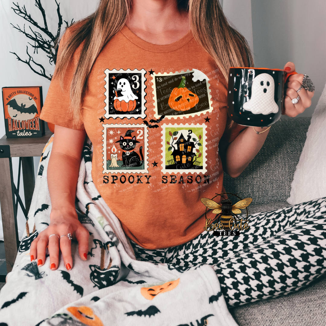 A woman wearing an orange crewneck t-shirt with a 'Spooky Season' graphic print featuring four Halloween-themed postage stamps. She is holding a black mug with a white ghost design and sitting on a couch next to a black-and-white houndstooth blanket.