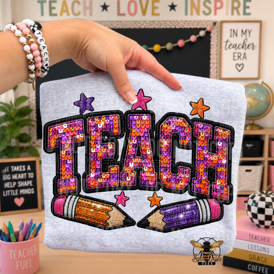 A folded light gray T-shirt is held by a hand, featuring a large sequined 'TEACH' graphic with two sequined pencils and small star accents. The background shows a styled classroom-themed room with chalkboard, signs, and desk decor.