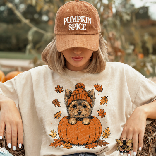 A woman wearing a tan baseball cap that says 'PUMPKIN SPICE' and a cream-colored graphic t-shirt featuring a small dog in an orange pumpkin with autumn leaves. She is posed outdoors in a pumpkin patch setting.