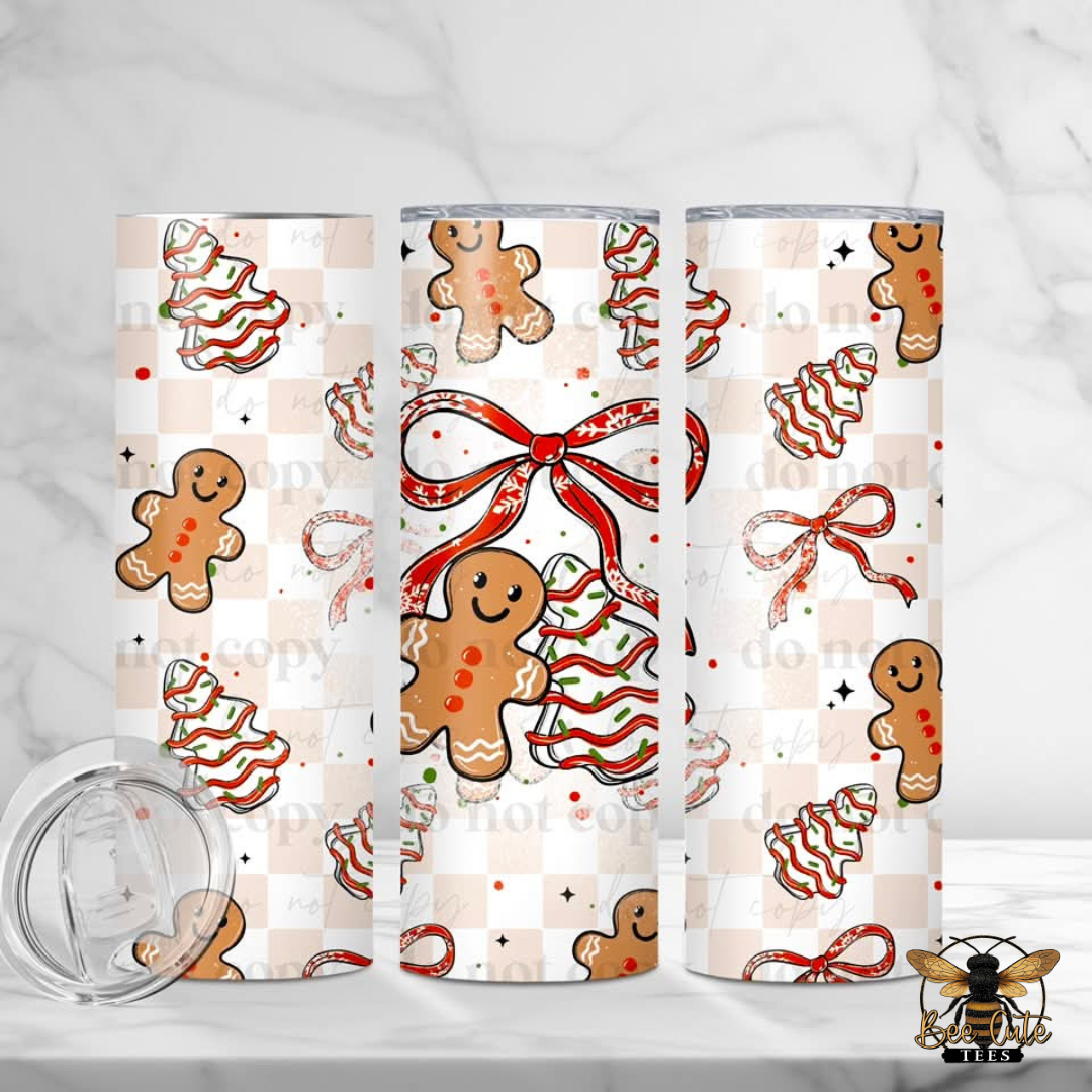 Three tall insulated tumblers with a Christmas gingerbread pattern, shown on a white marble surface with a clear lid lying in the foreground. The pattern features gingerbread men, gingerbread trees, and red bows on a beige and white checkered background.