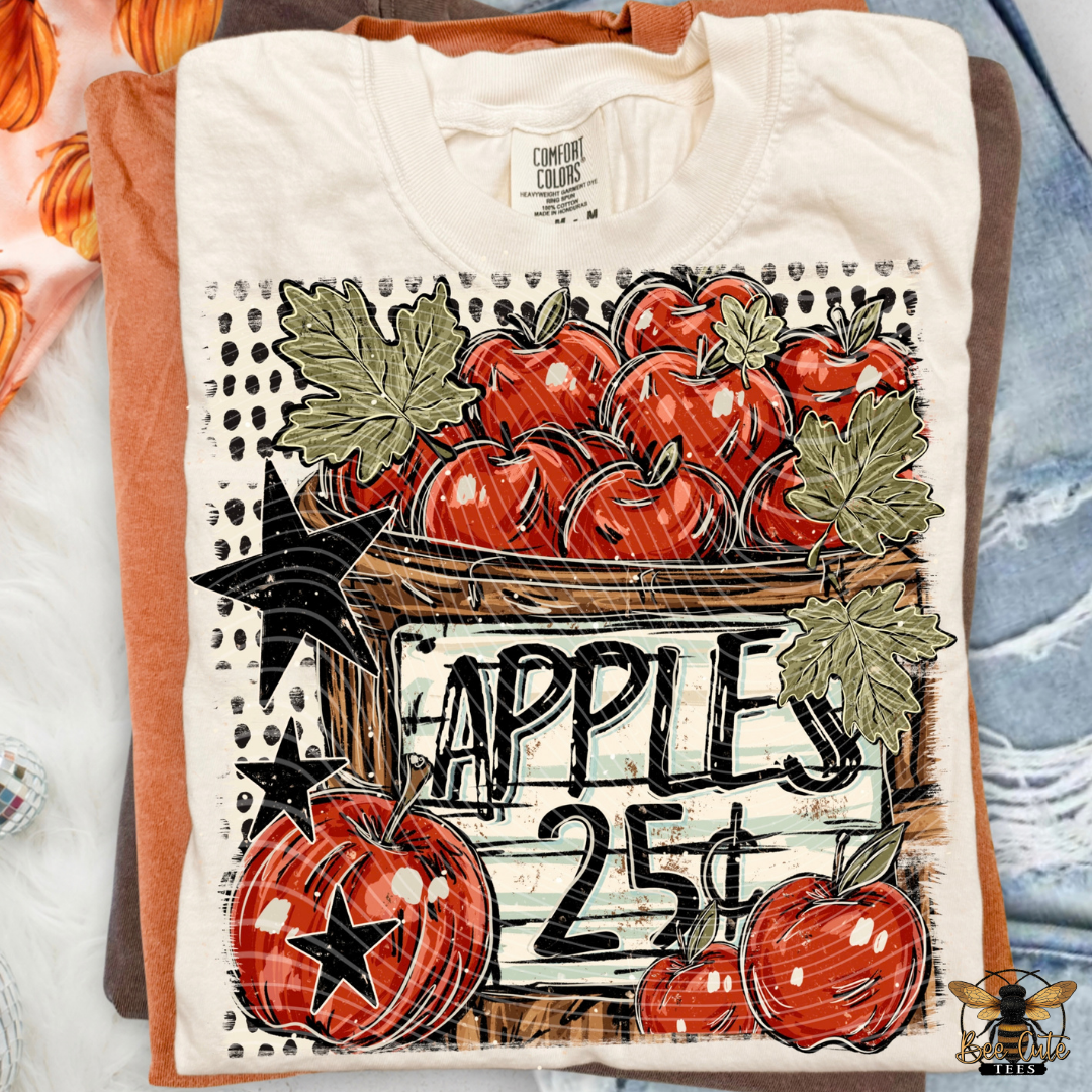 A folded cream-colored t-shirt with a vintage-style autumn apple graphic, displayed on top of orange and blue shirts. The graphic features red apples, green leaves, black stars, and a wooden crate sign reading 'APPLES 25¢'.