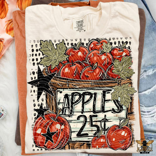A folded cream-colored t-shirt with a vintage-style autumn apple graphic, displayed on top of orange and blue shirts. The graphic features red apples, green leaves, black stars, and a wooden crate sign reading 'APPLES 25¢'.