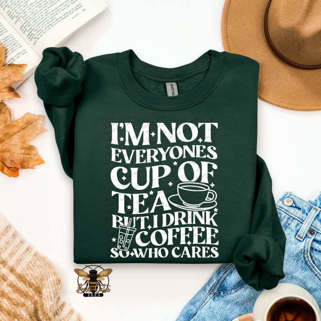 A dark forest green crewneck sweatshirt with a white graphic text print that reads 'I'M NOT EVERYONE'S CUP OF TEA BUT I DRINK COFFEE SO WHO CARES' accompanied by a coffee cup and iced coffee illustration. The sweatshirt is presented in a styled flat-lay arrangement with a tan hat, blue jeans, a book, autumn leaves, a knit sweater, and a cup of coffee on a white background.