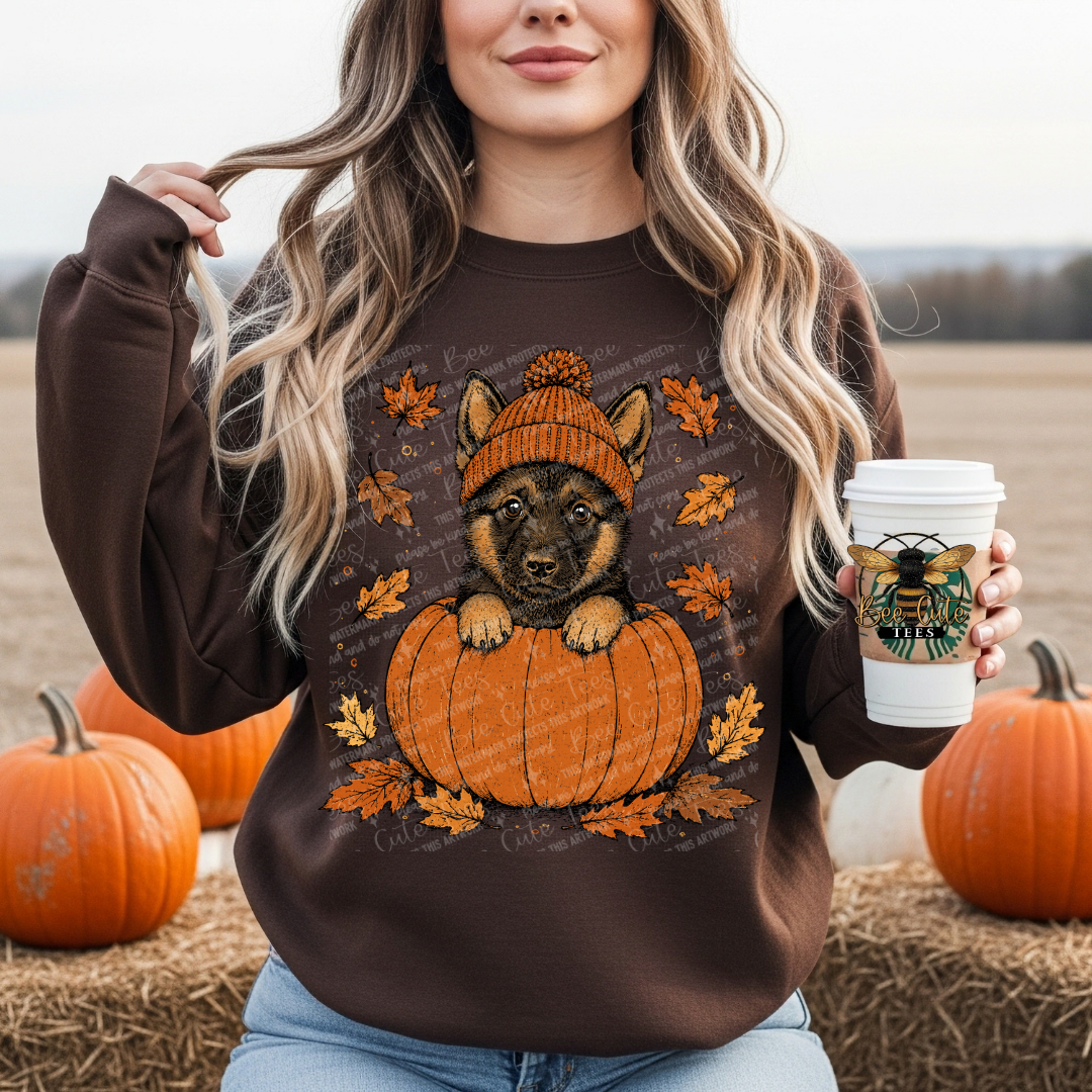 A woman wearing a brown crewneck sweatshirt with a graphic of a puppy in an orange knit hat sitting on an orange pumpkin surrounded by autumn leaves. She is holding a coffee cup and standing outdoors on a hay bale with pumpkins in the background.