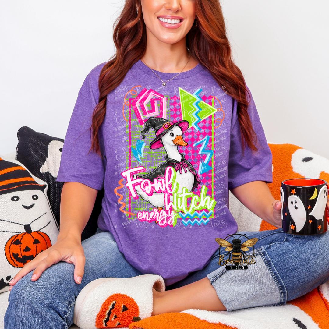 A woman wearing a purple graphic t-shirt with a duck and Halloween-themed design, sitting on an orange and white couch with Halloween pillows and a mug.