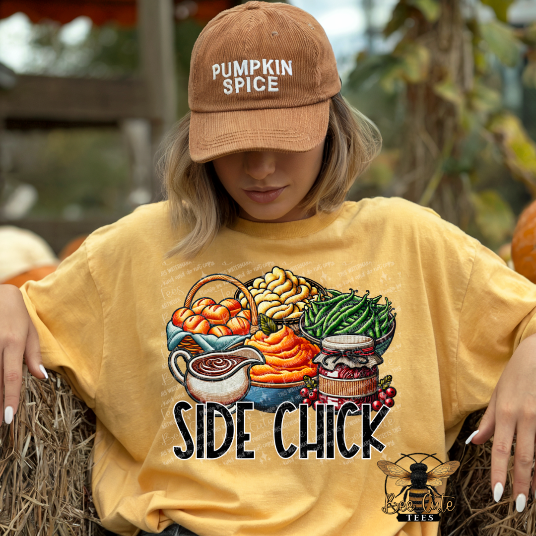 A woman wearing a tan baseball cap that says 'PUMPKIN SPICE' and a yellow graphic T-shirt with a Thanksgiving food illustration and the text 'SIDE CHICK'. She is sitting on a hay bale outdoors with pumpkins and autumn foliage in the background.