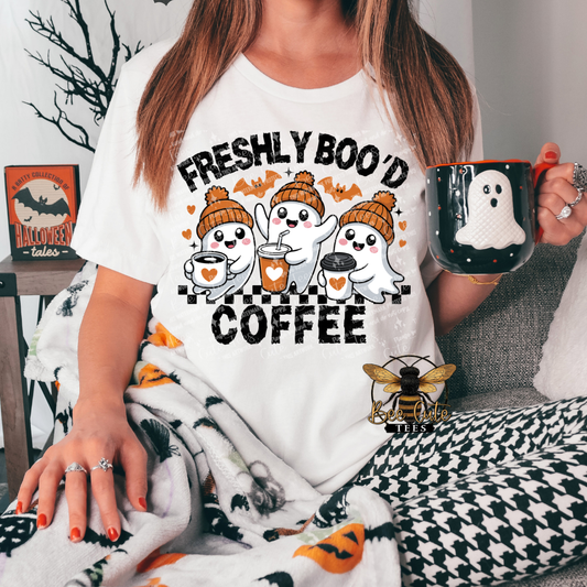 A woman wearing a white graphic t-shirt with a 'Freshly Boo'd Coffee' Halloween design, holding a black mug with a white ghost, while wrapped in a black and white houndstooth blanket with orange pumpkin accents.