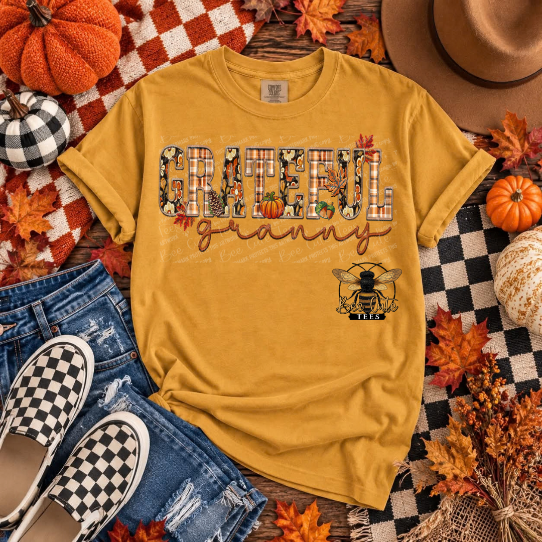 A mustard yellow graphic t-shirt laid flat on a rustic wood background with autumn-themed props including pumpkins, fall leaves, a cowboy hat, jeans, and slip-on shoes. The shirt features a 'Grateful Granny' design.