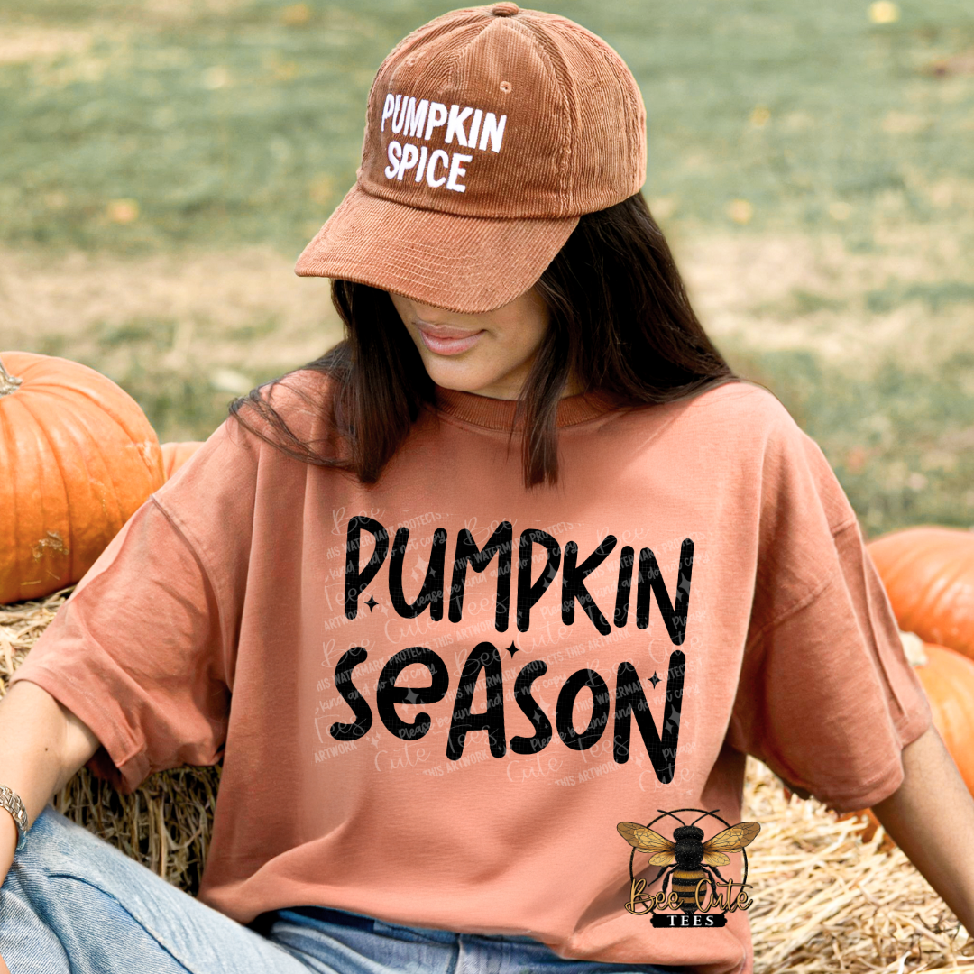 A woman sitting on a hay bale outdoors among pumpkins, wearing an oversized terracotta t-shirt with 'PUMPKIN SEASON' printed in black and a matching brown cap that says 'PUMPKIN SPICE'.