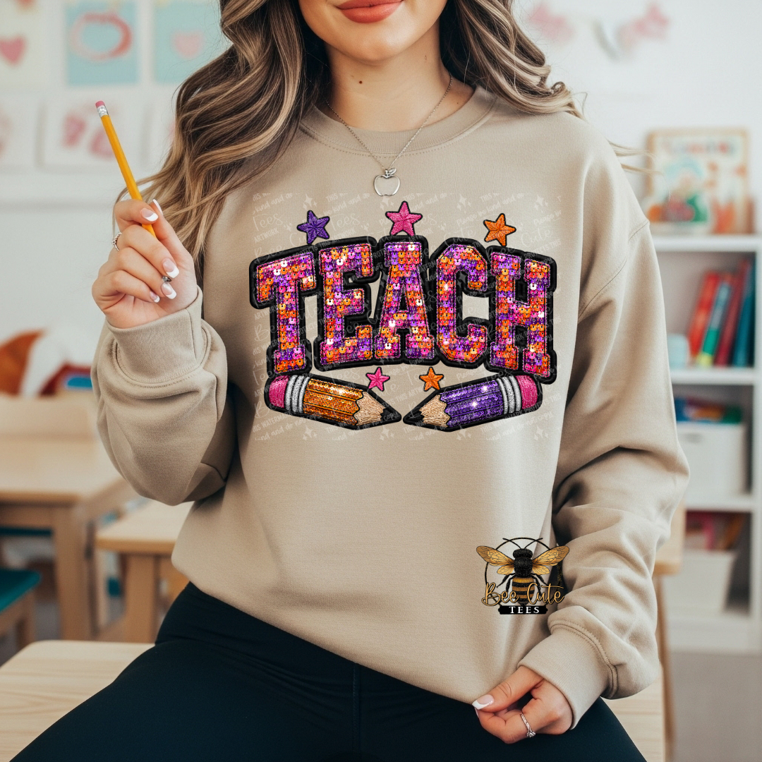 A woman wearing a beige crewneck sweatshirt with a colorful 'TEACH' graphic made of glitter-style letters, stars, and two pencils across the front. She is holding a yellow pencil in a classroom-like setting.