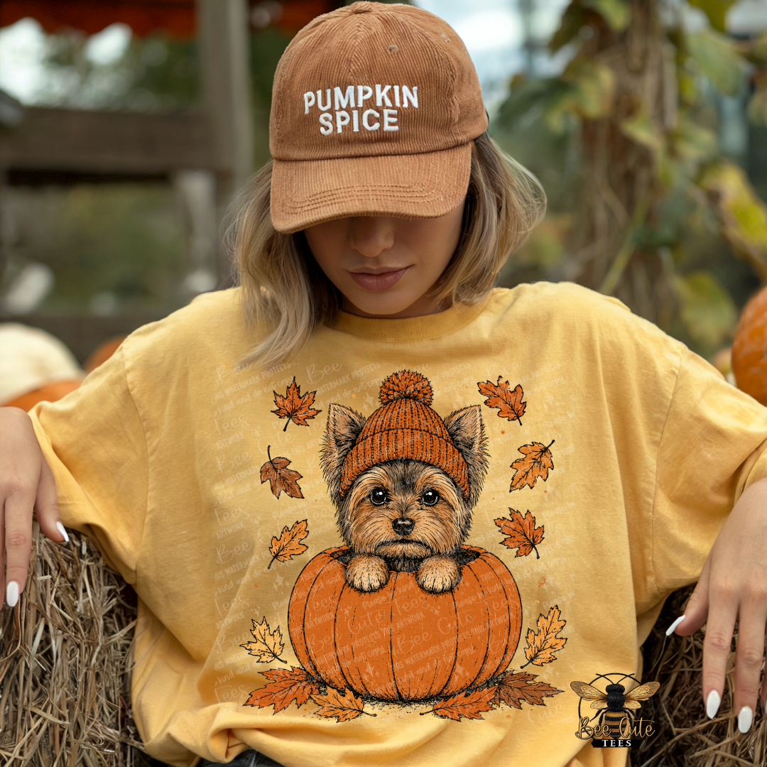 A woman wearing a tan 'Pumpkin Spice' baseball cap and a yellow graphic t-shirt with a cartoon dog in a pumpkin design, sitting on a hay bale at a pumpkin patch.