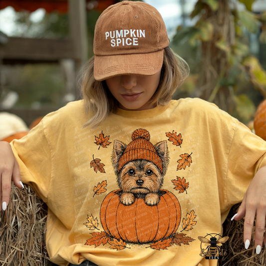 A woman wearing a tan 'Pumpkin Spice' baseball cap and a yellow graphic t-shirt with a cartoon dog in a pumpkin design, sitting on a hay bale at a pumpkin patch.
