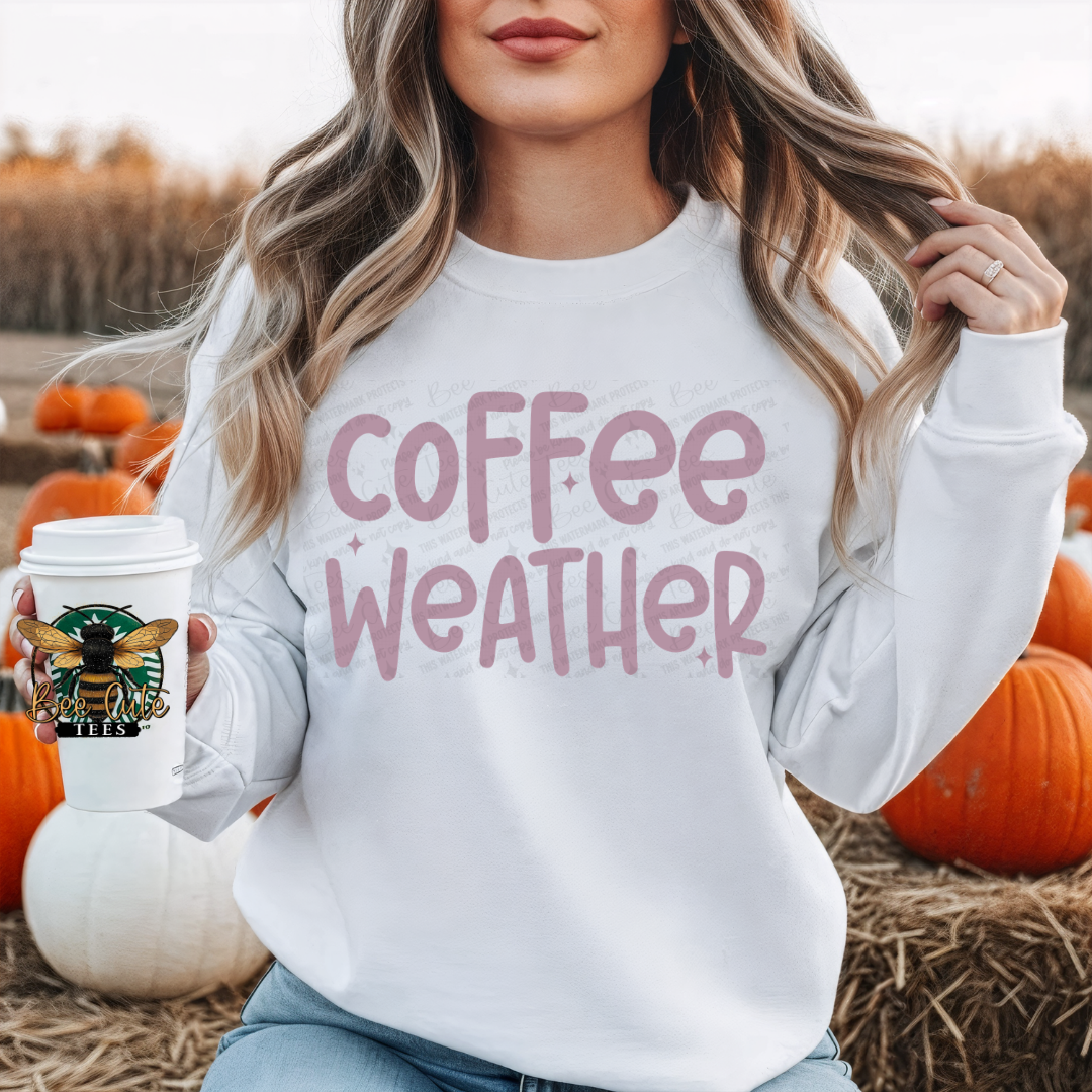 A woman in a white sweatshirt with a pink 'coffee weather' graphic holds a coffee cup in front of a pumpkin patch.