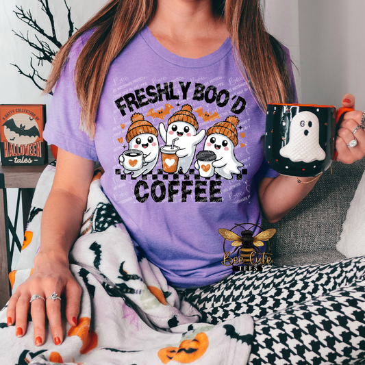 A woman wearing a purple graphic t-shirt that reads 'FRESHLY BOO'D COFFEE' with three cartoon ghosts wearing orange knit hats holding coffee cups. She is holding a black mug with a white ghost, wrapped in a black-and-white houndstooth blanket with orange pumpkin accents.