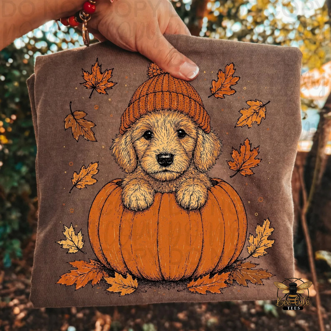 A hand holds a brown folded t-shirt, featuring a cartoon illustration of a puppy sitting on a pumpkin with orange autumn leaves. The background is a blurred outdoor forest scene.