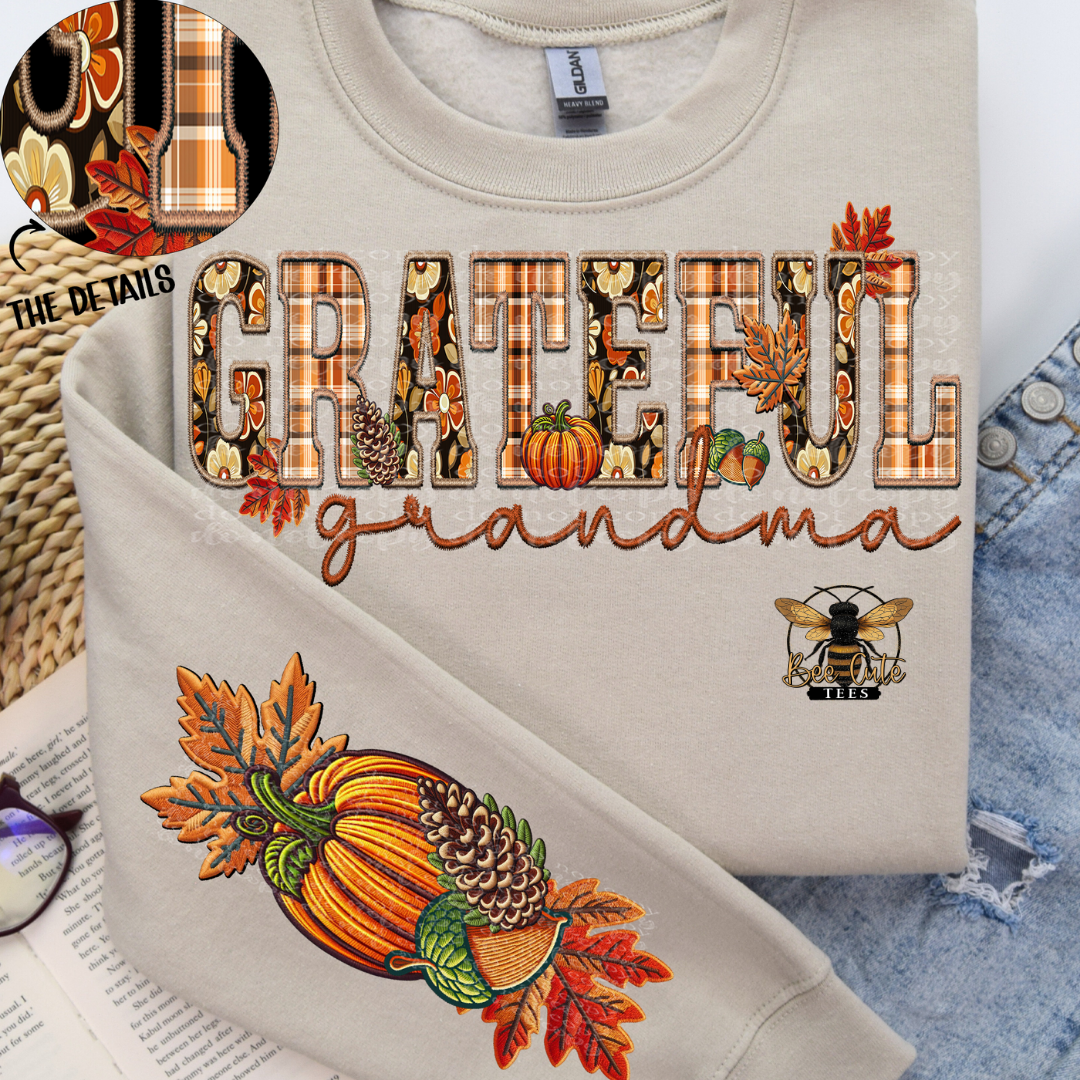 A folded light beige crewneck sweatshirt with a fall-themed graphic reading 'GRATEFUL grandma' in patterned letters, autumn leaves, pumpkins, and a bee logo. A circular inset shows fabric detail, and the sweatshirt is styled on top of denim with a book and glasses nearby.