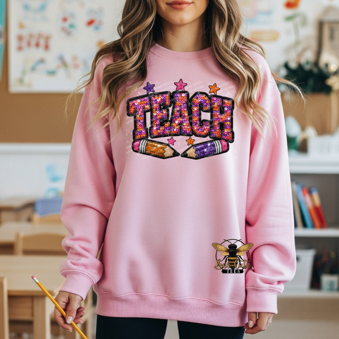A woman wearing a pink crewneck sweatshirt with a colorful 'TEACH' graphic made of glitter-style letters and two pencils, plus a bee patch on the lower right front. The background shows a softly blurred classroom interior.