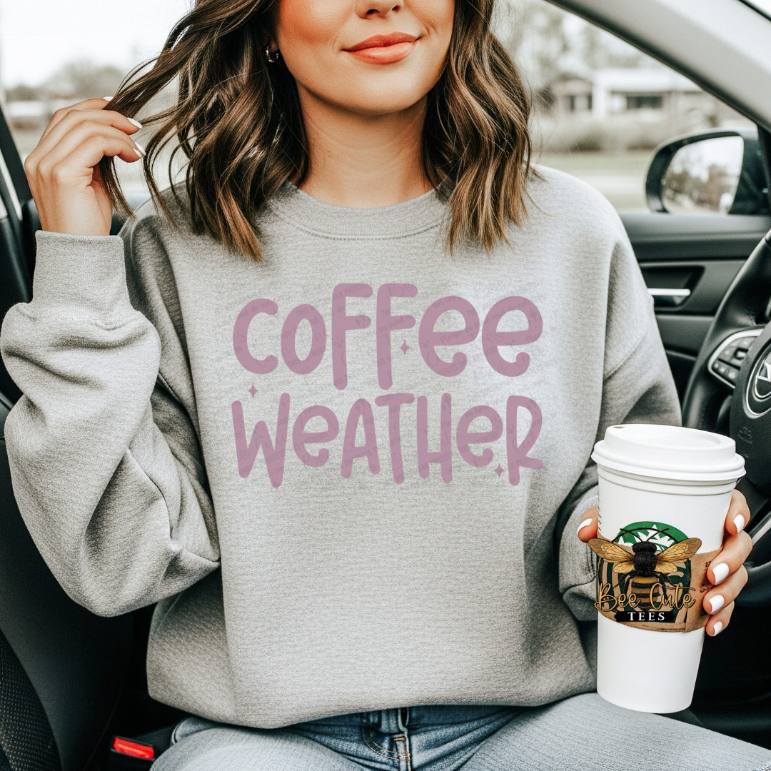 A woman sitting in a car wearing a light gray sweatshirt with the words 'coffee weather' printed in dusty pink. She is holding a white  coffee cup with a bee graphic.