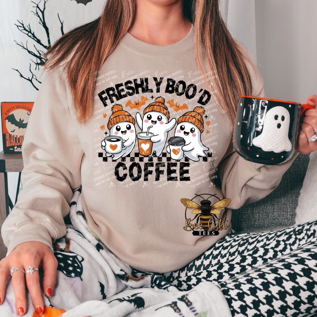 A woman wearing a beige crewneck sweatshirt with a Halloween graphic that reads 'FRESHLY BOO'D COFFEE' above three cartoon ghosts and below a small bee logo. She is holding a black mug with a white ghost design.