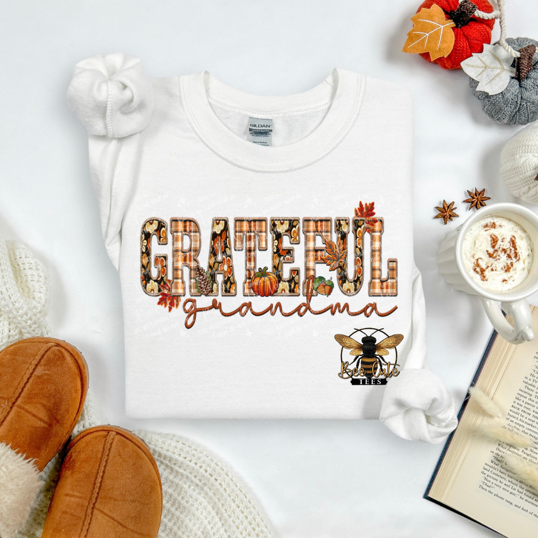 A white long-sleeve sweatshirt laid flat on a light background, featuring a fall-themed graphic that reads 'GRATEFUL grandma' with autumn leaves, pumpkins, and a bee. The sweatshirt is styled with cozy props including tan slippers, a mug of hot cocoa, an open book, and small seasonal decorations.