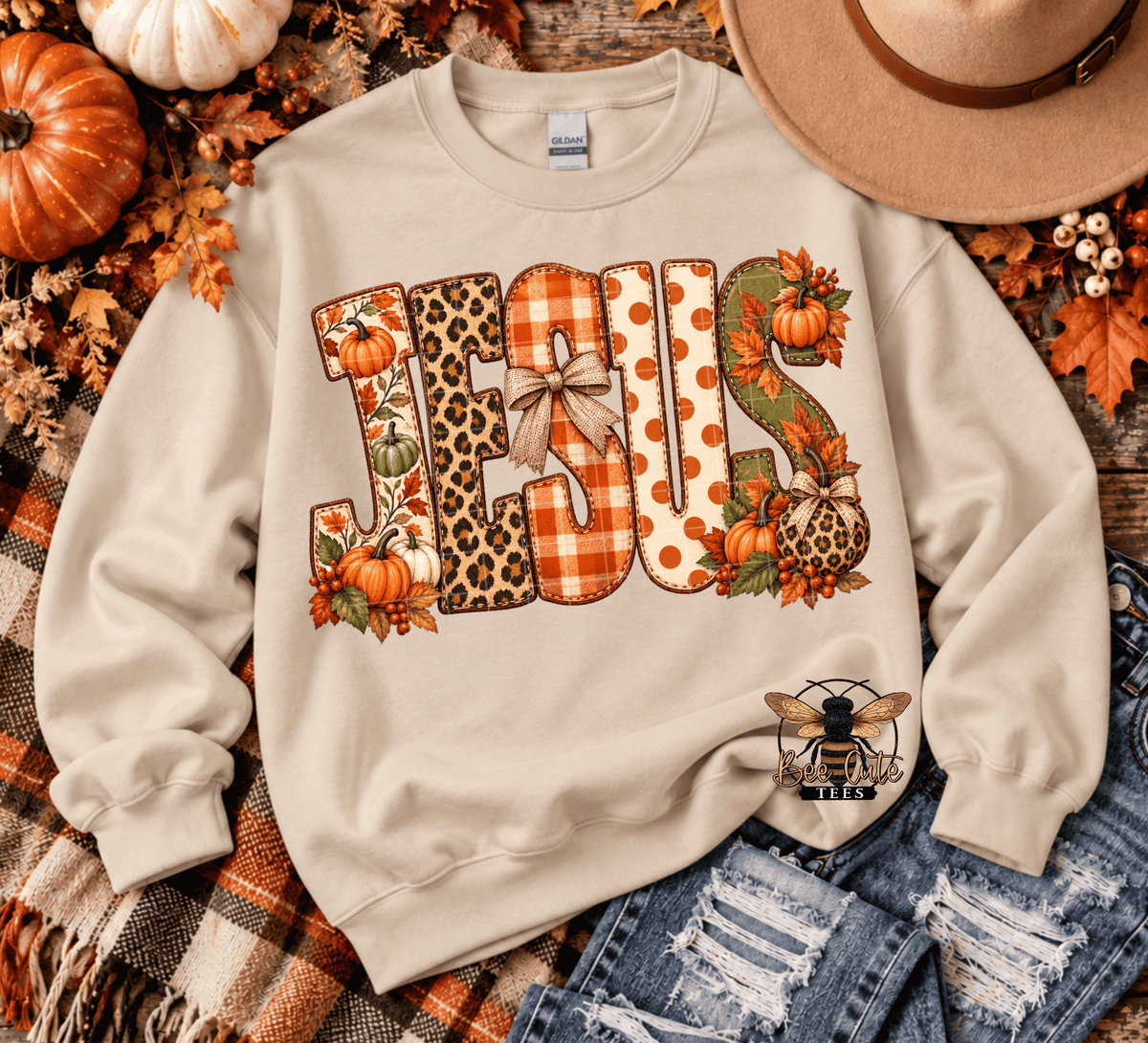 A cream-colored crewneck sweatshirt featuring a large 'JESUS' graphic in autumn-themed patterns (plaid, leopard print, polka dots) with pumpkins and leaves. The sweatshirt is styled in a flat lay with a plaid blanket, distressed denim, a tan hat, and pumpkins.